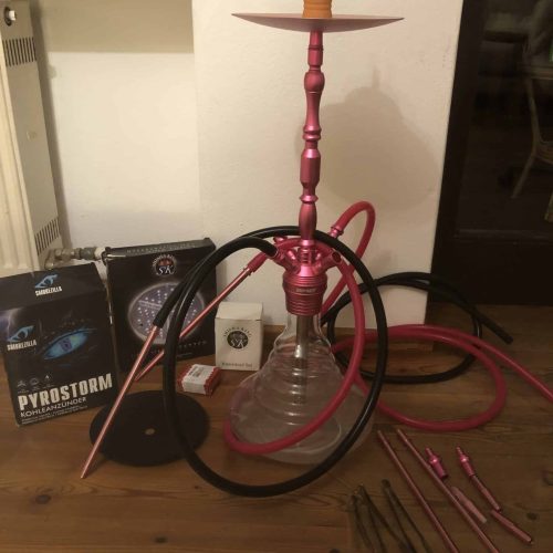 Shisha mit pinkem Metallrahmen und Glasbowl, Zubehör und Tabak im Hintergrund.