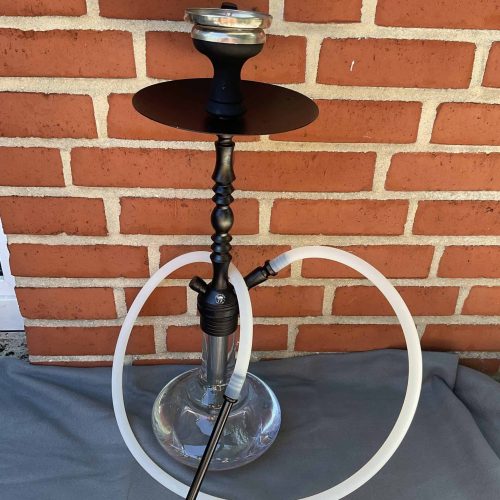 Shisha mit Glasbowl, Schlauch und Metallstück vor Ziegelwand.
