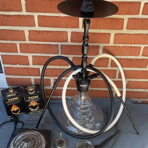 Shisha mit Glasbowl, Heizplatte und Zubehör vor Ziegelwand.