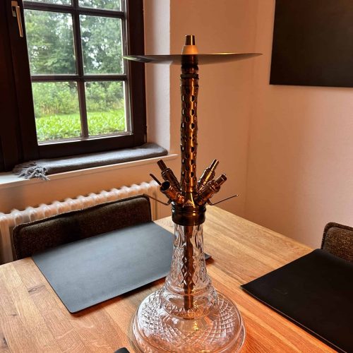 Glasbowl Shisha mit Metallaufsatz auf Holztisch neben Fenster.