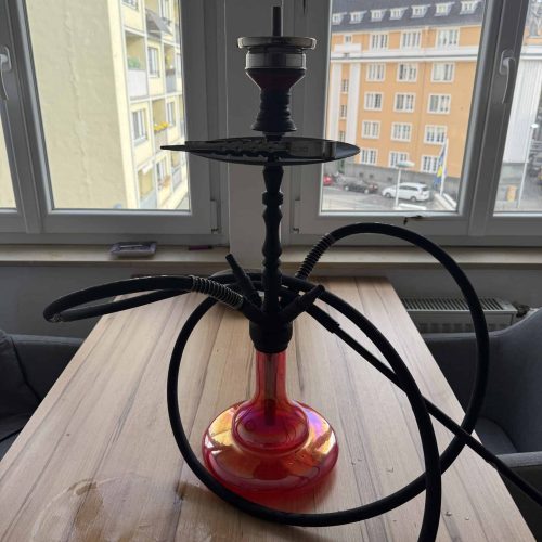 Shisha mit schwarzem Schlauch und rotem Glasboden, Blick auf Stadt im Hintergrund.