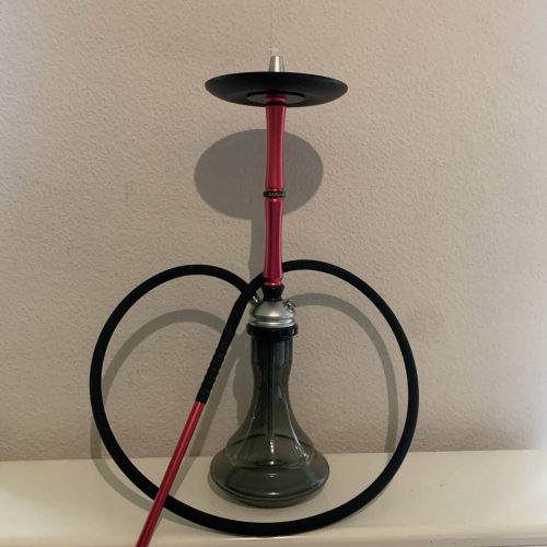 Shisha mit rotem Schlauch, Glasbowl und Metallaufsatz, auf weißem Tisch gegen Wand.