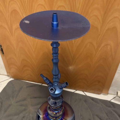 Hookah mit Glasbasis und Metallrohr, auf Teppich vor Holzdecke.