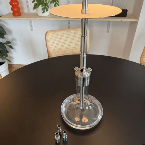 Moderne Glas-Shisha mit Metallaufsatz auf schwarzem Tisch, Zubehör im Vordergrund.