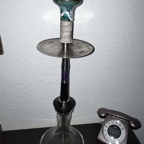 Wasserpfeife mit Glasbasis und Metallaufsatz, auf Tisch mit Telefon im Hintergrund.