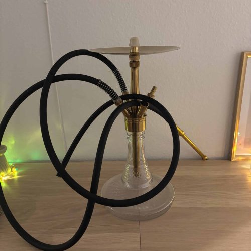 Shisha mit Glasbasis und Metallaufsatz, auf einem Holztisch, dekorativ beleuchtet.