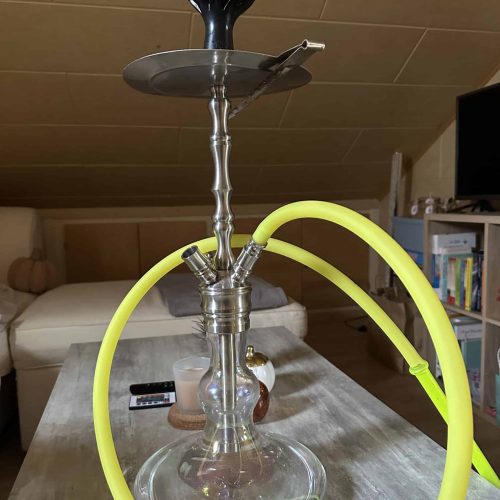 Shisha mit Glasbasis und Metallaufsatz, gelber Schlauch, auf Tisch im Wohnzimmer.