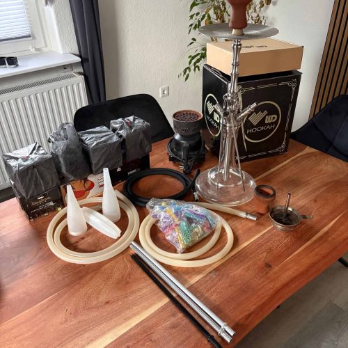 Shisha mit Glas- und Metallteilen, Tabak, Kohle und Zubehör auf Tisch.