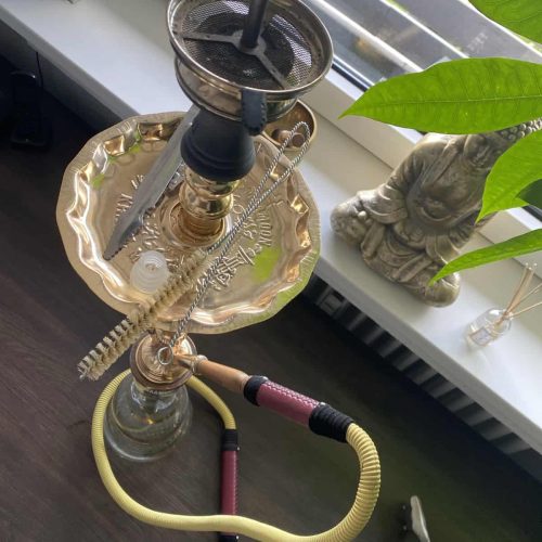 Shisha mit Glasbowl, Schlauch und Tabak, auf dunklem Tisch neben Fenster und Dekofigur.