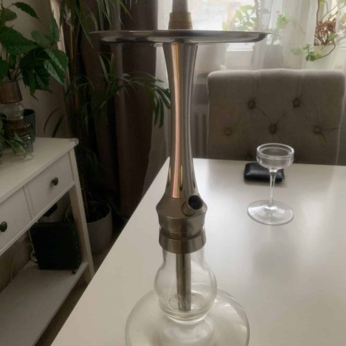 Wasserpfeife mit Metallaufsatz auf weißem Tisch, Wohnzimmer im Hintergrund.