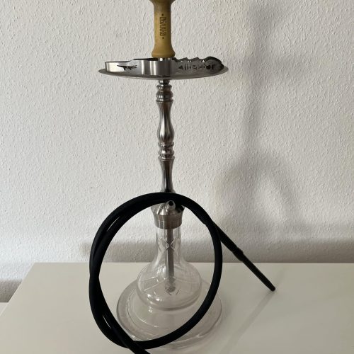 Rauchsäule mit Shisha, Glasbowl, Metallrohr und Schlauch, auf weißem Tisch gegen weiße Wand.
