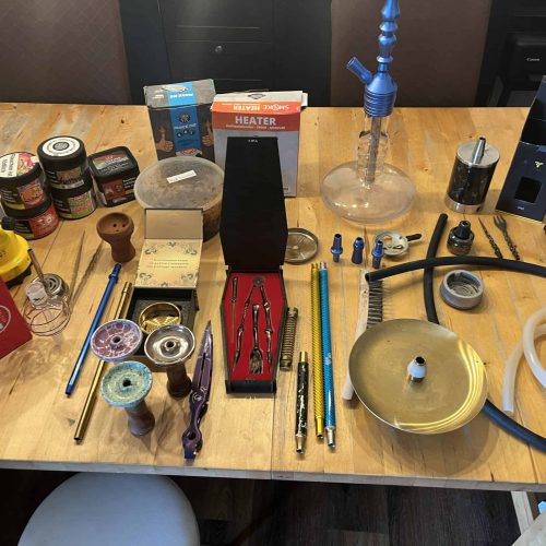 Verschiedene Shisha-Teile, Glasbowl, Schlauch, Zange und Zubehör auf Tisch.