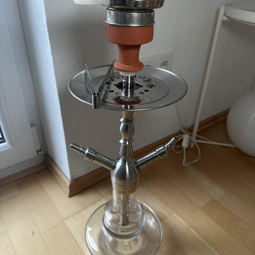Rauchsäule mit Shisha-Setup, bestehend aus Glas, Metall und Silikon, auf Holzboden neben Fenster.