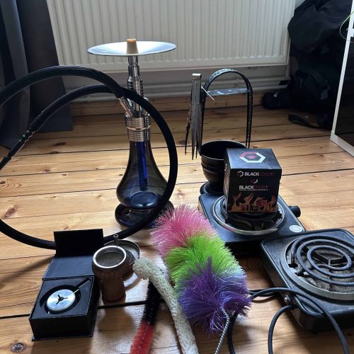 Shisha mit Glasbowl, Schlauch und Kohle, auf Holzboden in Raum.