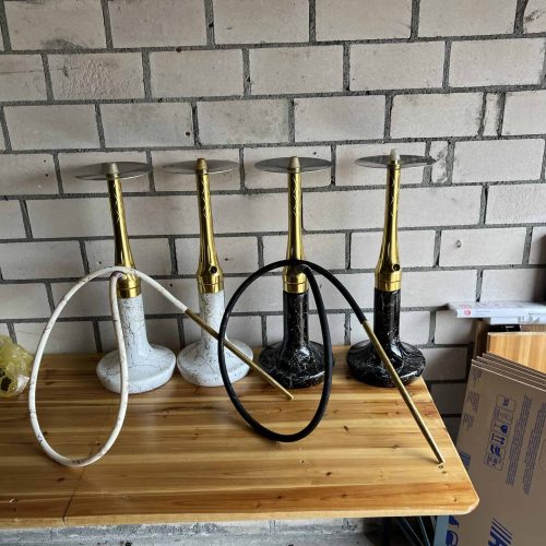 Verschiedene Shisha-Hooks in Schwarz und Weiß auf Holztablett, Ziegelwand im Hintergrund.