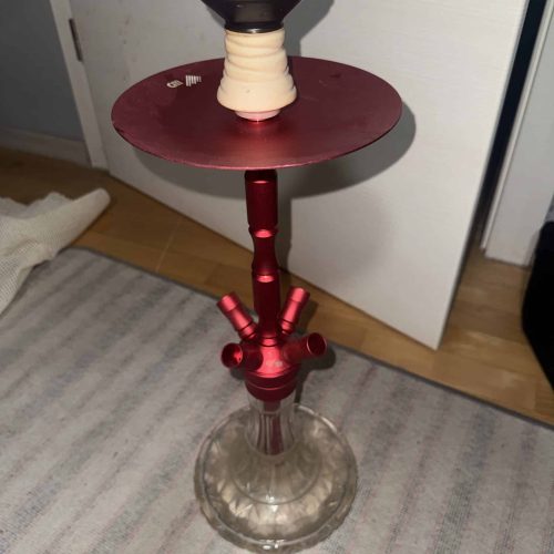 Shisha mit Glasbowl, Metallstücken und Schlauch, auf Boden stehend.