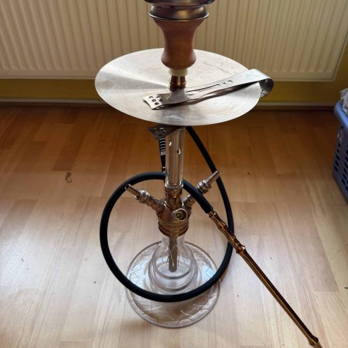 Shisha mit Glasbowl, Tonkopf und Metallzubehör auf Tisch.