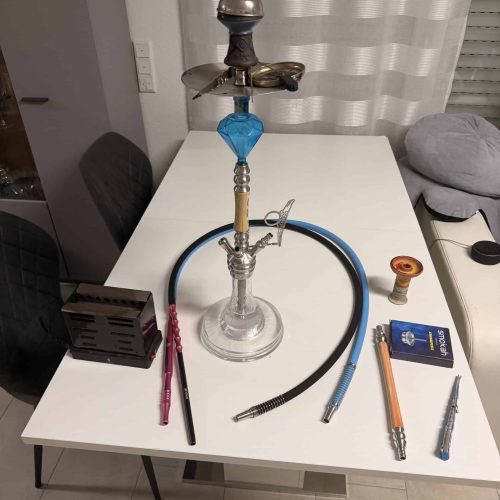 Shisha mit Glasbowl, Schlauch und Zubehör auf Tisch.