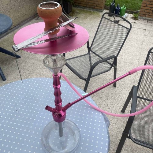 Shisha mit Glasbowl auf Tisch im Garten, mit Zubehör im Hintergrund.