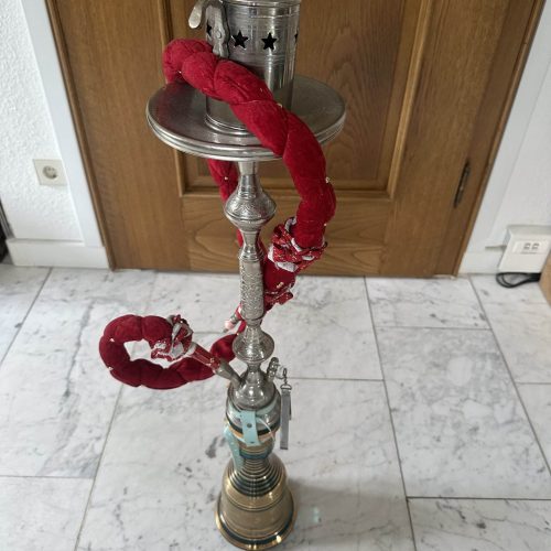 Rauch-Hookah mit silbernem Kopf, rotem Schlauch und Edelstahl-Design, steht auf Fliesen vor Holztür.