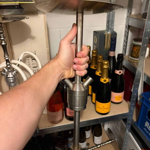 Hand hält eine Shisha-Pressvorrichtung in einem Lagerraum mit Flaschen im Hintergrund.