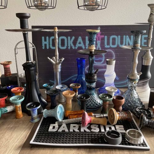 Verschiedene Wasserpfeifen und Rauchschwerter auf einem Tisch, im Hintergrund ein Neon-Schild "Hooka.