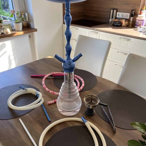 Shisha mit blauer Rauchsäule, Kopf und Schlauch auf Esstisch in Küche.