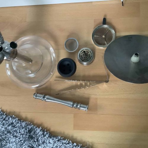 Shisha-Set mit Glasbowl, Metallteilen und Zubehör auf Holzboden.