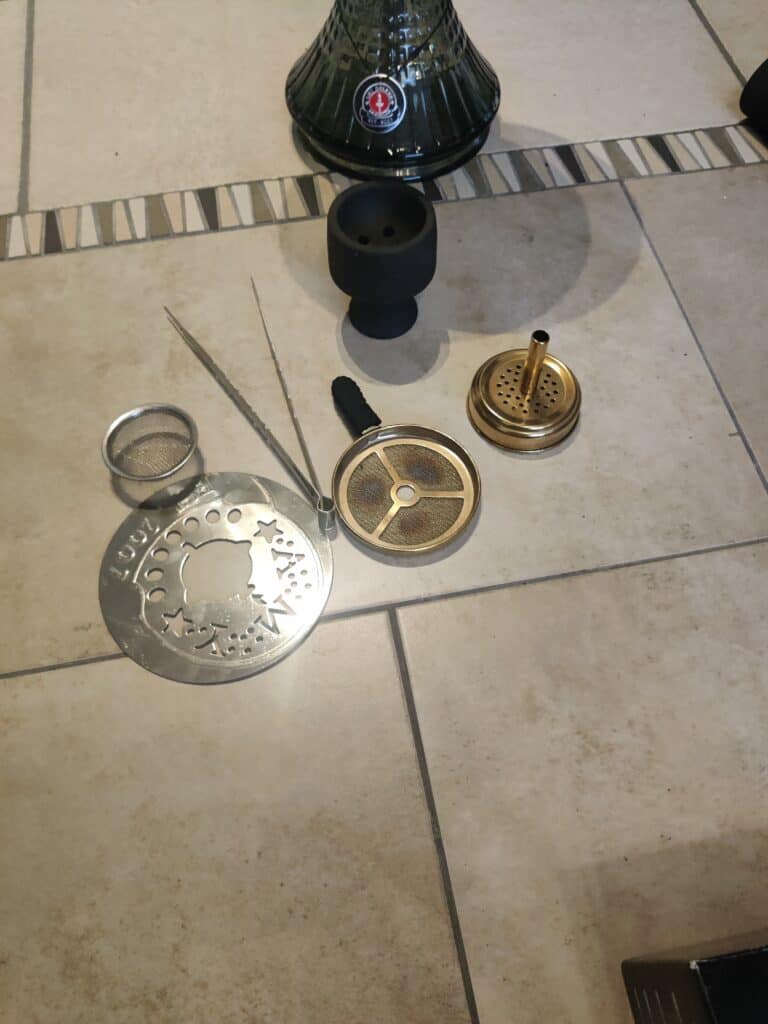 Verschiedene Shisha-Teile auf Fliesenboden, inklusive Glasbowl und Dichtungen.