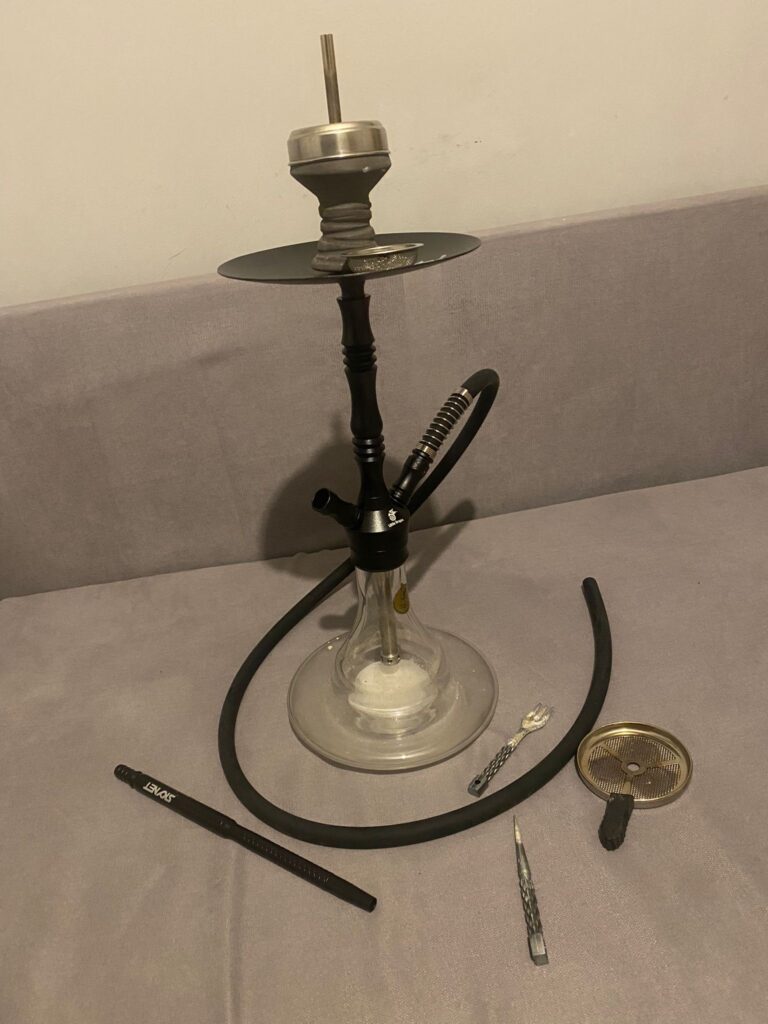 Shisha mit Glasbowl, Schlauch und Zubehör auf Tisch.