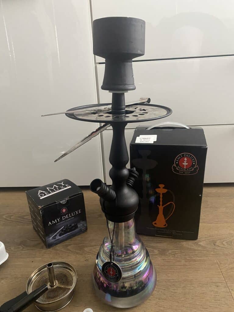 Shisha mit Glaskolben, Rauchsäule und Zubehör, auf Holztisch.